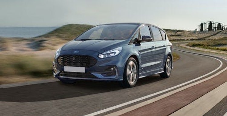 A blue car driving on a winding road near a coastline, with a clear sky and scenic landscape in the background.