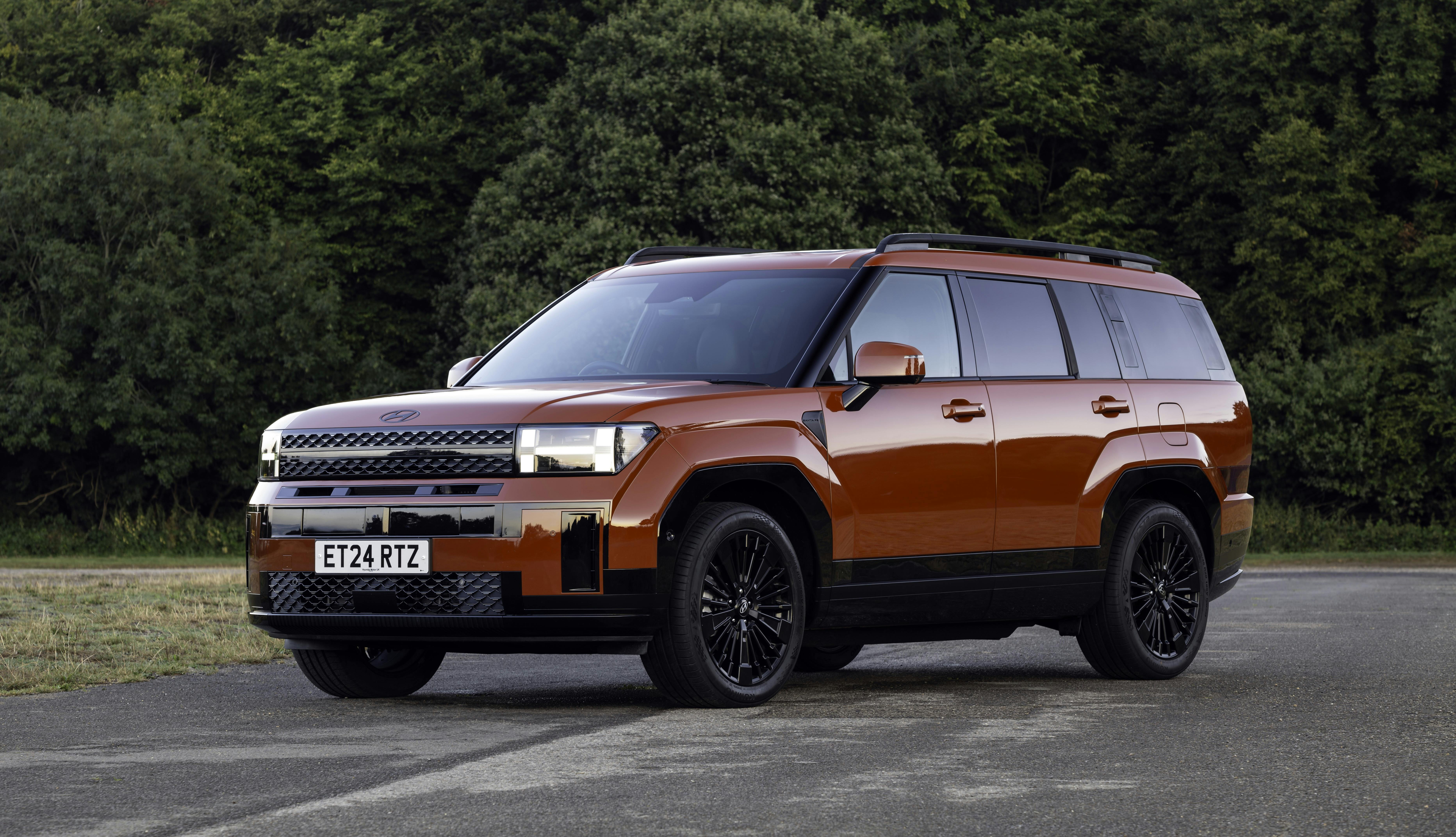 Red Hyundai Santa Fe parked on tarmac in front of trees, front view.
